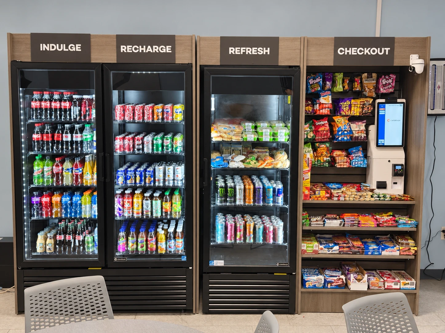 Break room micro market with drink coolers labeled Indulge, Recharge, and Refresh, a snack wall, and a self-checkout kiosk