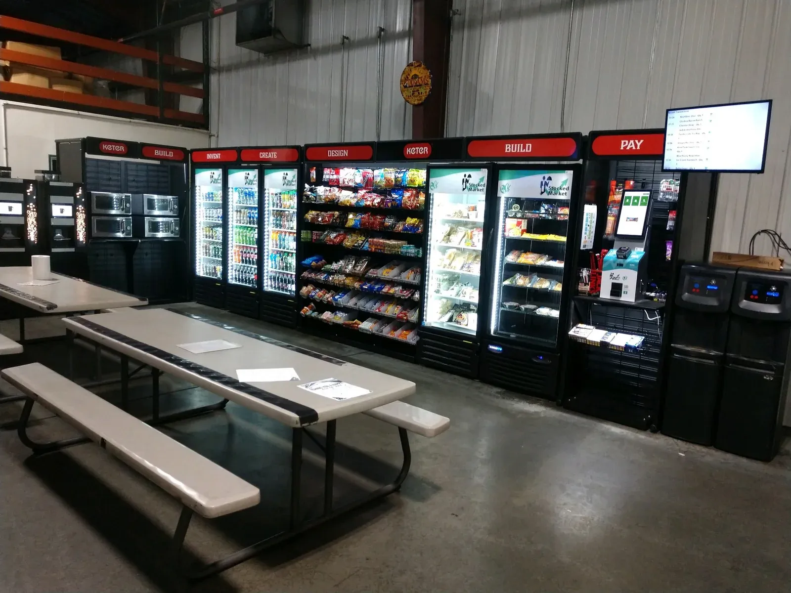 Warehouse break room micro market with drink coolers, snack racks, and a self-checkout kiosk beside picnic tables