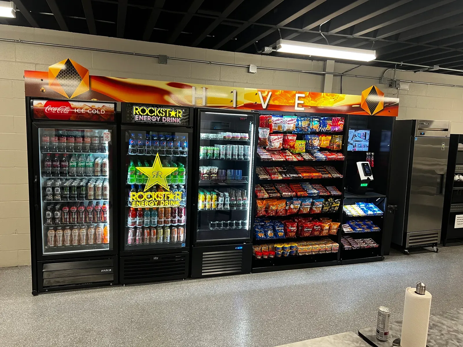 Micro market wall with glass-door drink coolers, an open snack rack, and a self-checkout kiosk