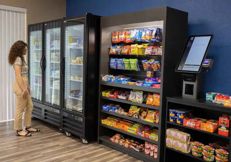 Woman shopping a micro market with coolers, snack shelves, and a self-checkout kiosk